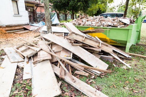 A pile of discarded wooden boards and planks, featuring weathered surfaces and varying lengths, is scattered on a grassy area near a green skip filled with rubble and broken bricks. The planks show signs of age with some chipped edges and a rough texture, while the bricks in the skip are irregular in shape and in shades of red and brown. In the background, there is a residential building with a small set of stairs and some trees providing partial shade, indicative of a suburban outdoor setting in the vicinity of Feltham. The scene appears to be part of a waste clearance process conducted by Commercial Waste Removal Feltham, with the arrangement suggesting ongoing rubbish removal or clearance work in a garden or driveway area.