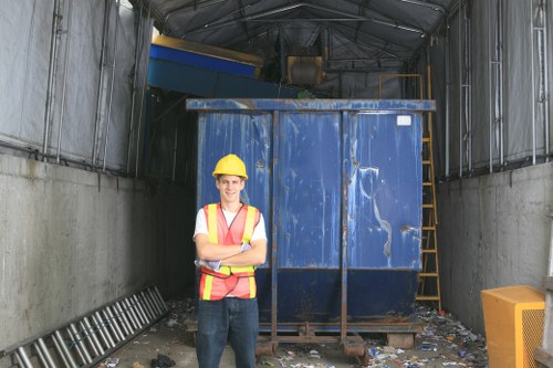An outdoor industrial waste storage area with a tall, blue metal skip situated on a concrete surface, surrounded by grey corrugated metal walls on either side. The skip has signs of wear including scratches and faded paint, and is positioned at the back of the space. To the right of the skip, there is a yellow ladder leaning against a supporting beam, and scattered rubbish and debris are visible on the ground. In the foreground, a male worker wearing a yellow safety helmet, a high-visibility vest with orange and yellow reflective stripes, a white shirt, and dark trousers is standing with his arms crossed, smiling at the camera. The setting is part of a logistics or waste management site, possibly near a commercial or industrial area in Feltham, with ambient lighting indicating daytime.