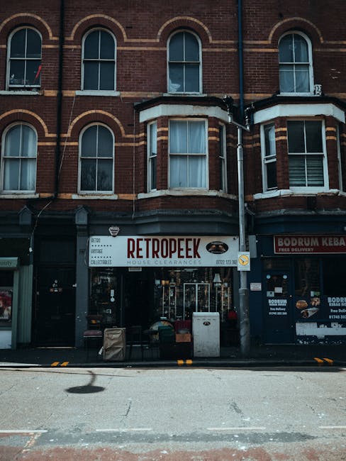 A busy urban street scene featuring a mix of commercial and retail buildings on Feltham High Street. In the foreground, a group of pedestrians walk along the pavement, some carrying shopping bags and 